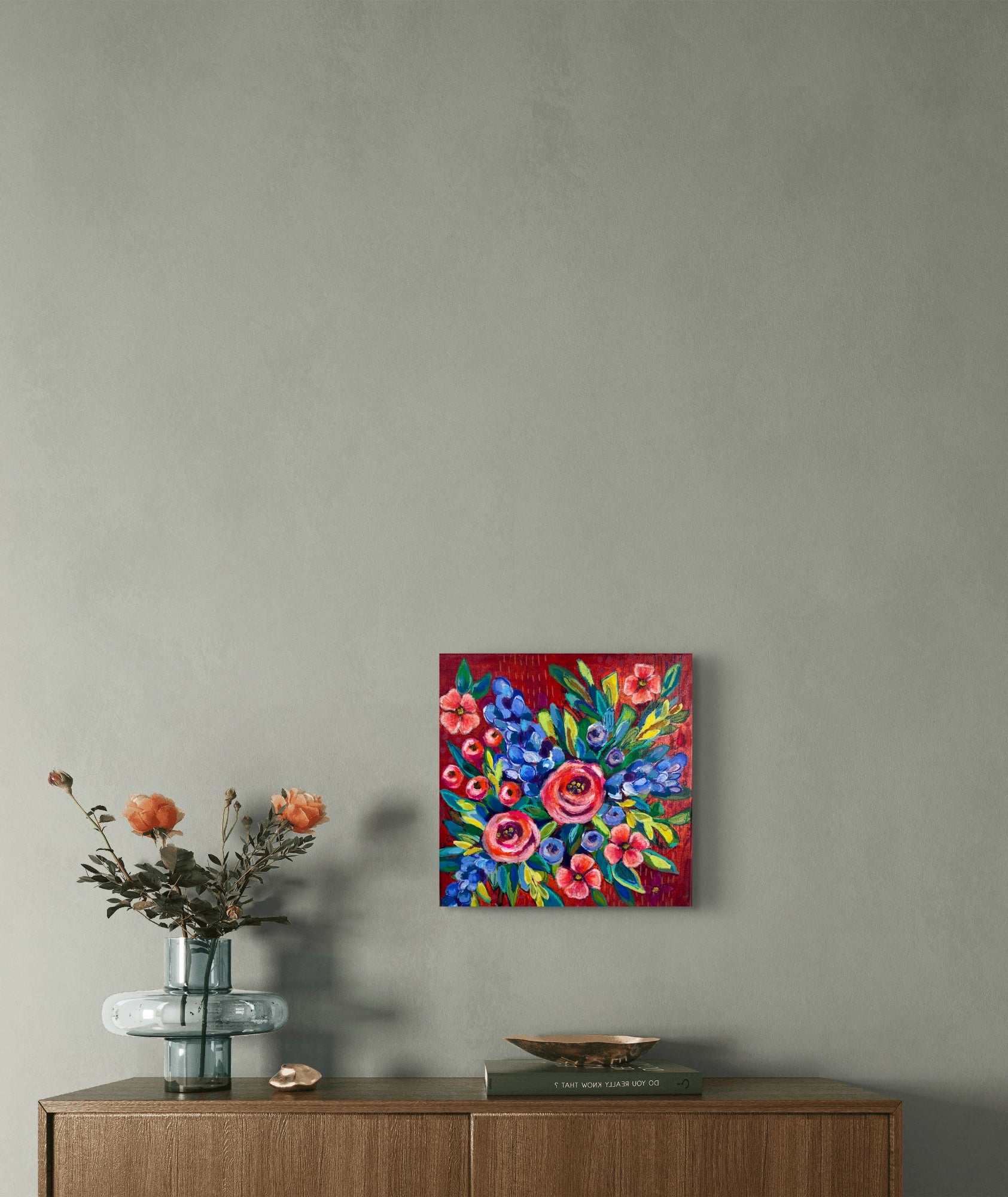 Colorful floral painting on a gray wall above a wooden cabinet with a vase and decorative items.